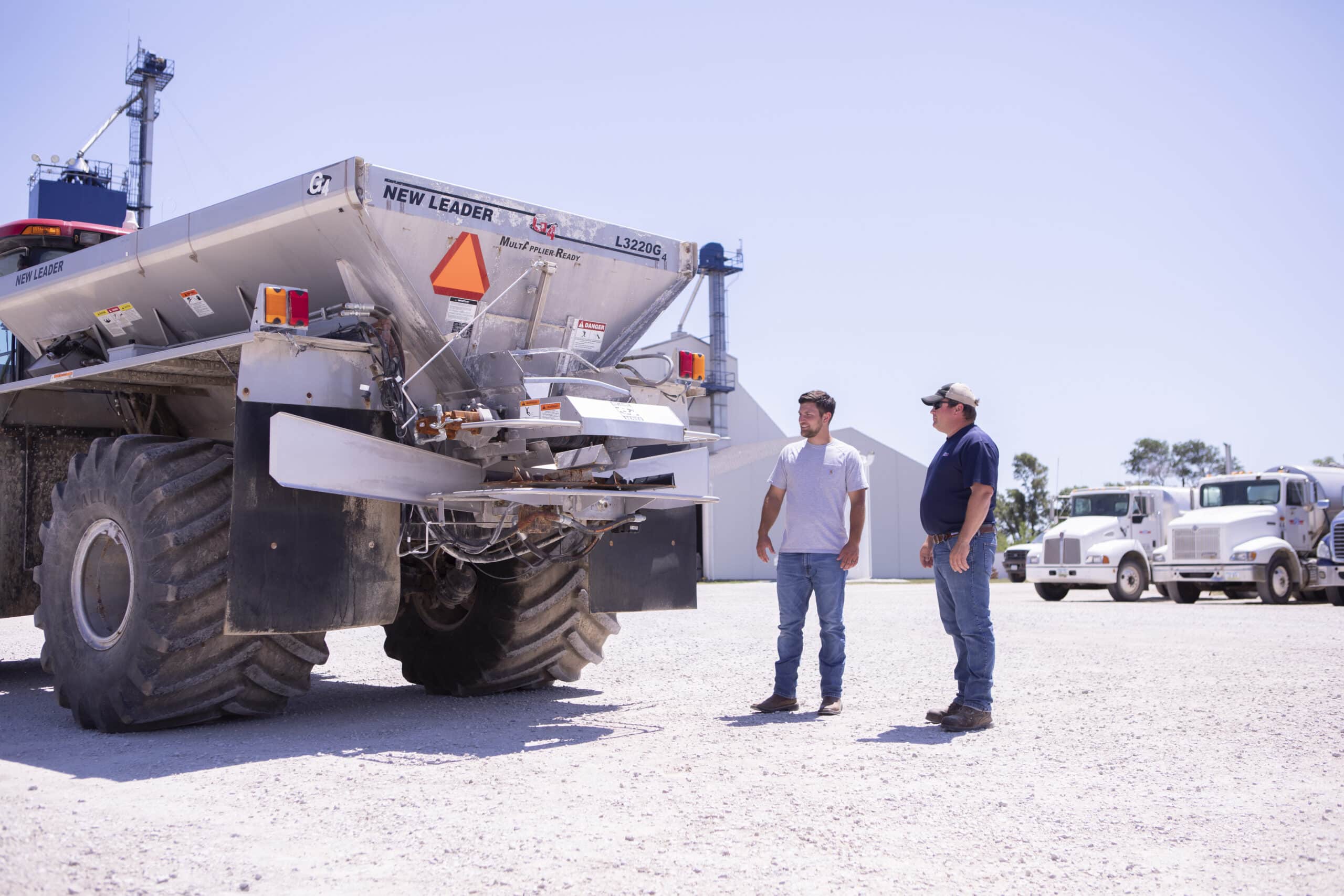Two men having a conversation while standing near the back of a fertilizer spreader.